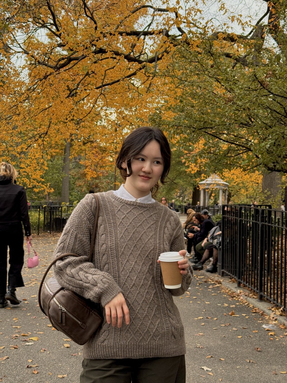 Jing in a hand-knit cable sweater, holding a coffee in an autumn park